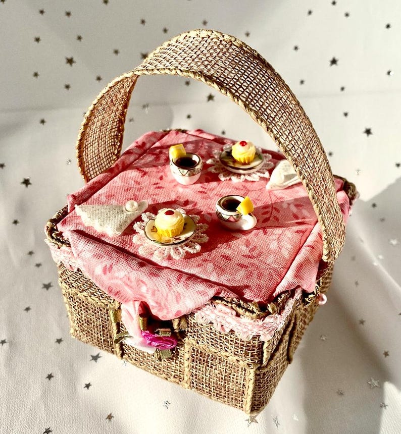May include: A miniature picnic basket with a burlap handle and a pink fabric lining. The basket is filled with miniature teacups, saucers, and cupcakes.
