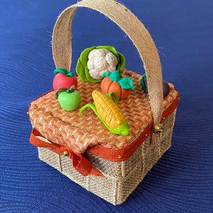 Pode incluir: Uma pequena cesta tecida de cor castanho com uma pega de corda. A cesta est&aacute; cheia de miniaturas de vegetais de barro, incluindo uma ma&ccedil;&atilde; verde, uma ma&ccedil;&atilde; vermelha, uma espiga de milho, uma ab&oacute;bora, uma couve-flor e um piment&atilde;o verde.