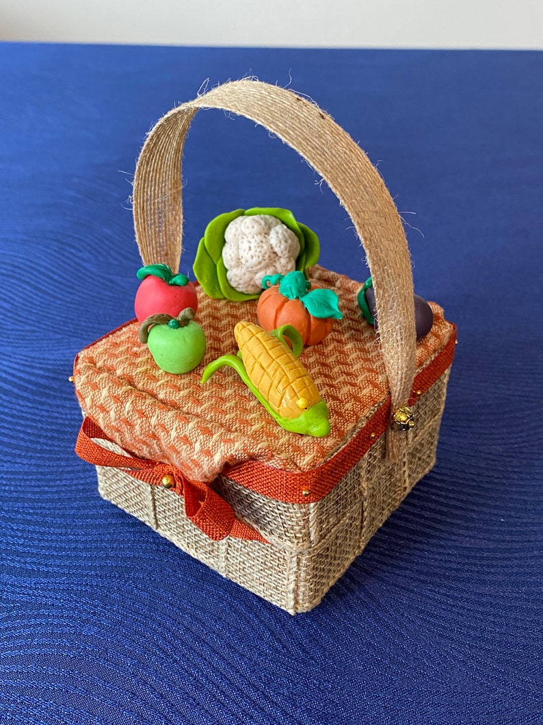 Pode incluir: Uma pequena cesta tecida de cor castanho com uma pega de corda. A cesta est&aacute; cheia de miniaturas de vegetais de barro, incluindo uma ma&ccedil;&atilde; verde, uma ma&ccedil;&atilde; vermelha, uma espiga de milho, uma ab&oacute;bora, uma couve-flor e um piment&atilde;o verde.