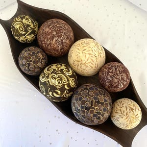 May include: A dark brown, elongated wooden bowl containing eight decorative spheres. The spheres feature various patterns including gold swirls, dark brown circles, and light beige leaf prints, arranged in a cluster within the curved vessel.