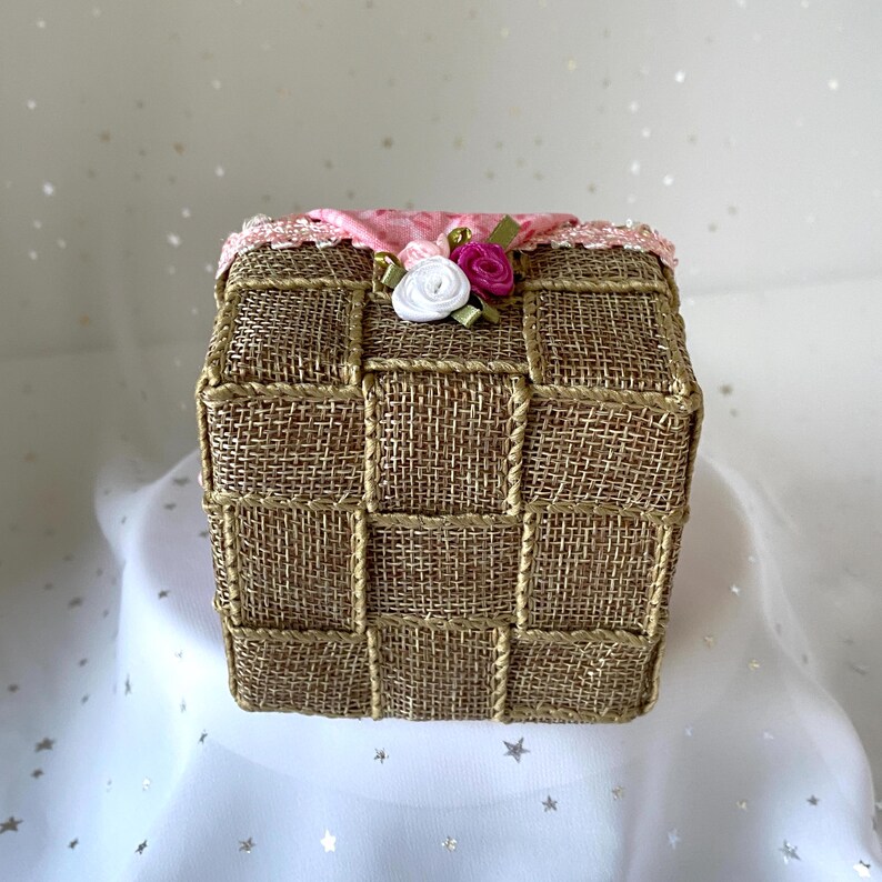 May include: A square tissue box cover made of woven burlap with a pink and white fabric trim and pink and white fabric flowers.