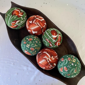 May include: Six fabric-covered decorative spheres arranged in a dark brown wooden bowl. Three spheres feature a green background with white football field markings and brown footballs, while three feature a pattern of brown footballs on red.