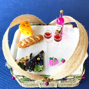 Pode incluir: Uma cesta de piquenique de vime em miniatura com um forro de tecido castanho e verde. A cesta cont&eacute;m um p&atilde;o, uma fatia de queijo, dois copos de vinho, uma garrafa de vinho com o r&oacute;tulo "Chateau Rose" e duas pequenas cachos de uvas cor-de-rosa.