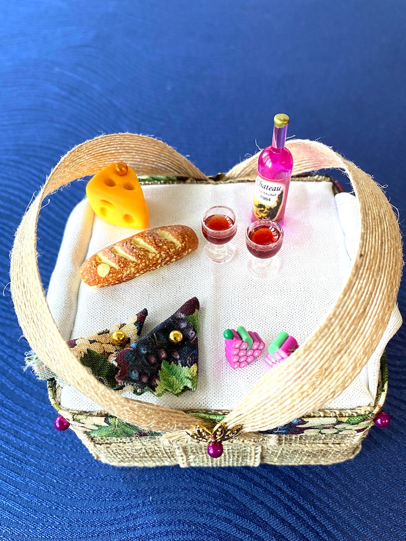 Pode incluir: Uma cesta de piquenique de vime em miniatura com um forro de tecido castanho e verde. A cesta cont&eacute;m um p&atilde;o, uma fatia de queijo, dois copos de vinho, uma garrafa de vinho com o r&oacute;tulo "Chateau Rose" e duas pequenas cachos de uvas cor-de-rosa.