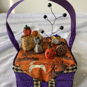 Pode incluir: Uma caixa roxa brilhante com uma borda xadrez preta e dourada e uma al&ccedil;a de fita roxa. A caixa &eacute; decorada com um tema de Halloween, incluindo um gato preto, uma coruja, ab&oacute;boras e uma lua crescente.
