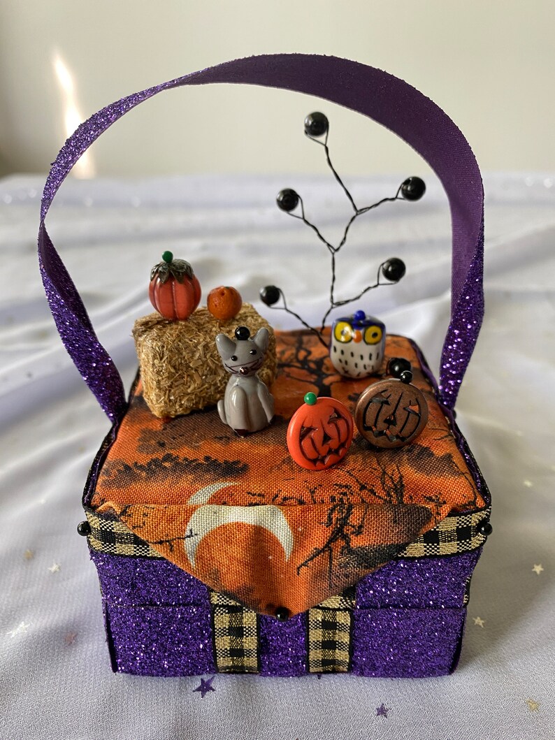 Pode incluir: Uma caixa roxa brilhante com uma borda xadrez preta e dourada e uma al&ccedil;a de fita roxa. A caixa &eacute; decorada com um tema de Halloween, incluindo um gato preto, uma coruja, ab&oacute;boras e uma lua crescente.