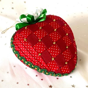 May include: A handmade strawberry ornament with a woven red and white polka dot design. The ornament is accented with gold beads, a green and white bow, and a gold hanging loop. The edges are trimmed with green and gold trim.