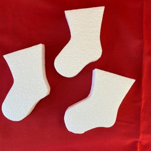 May include: Three white foam Christmas stocking cutouts on a red background. The stockings are textured and have a simple, classic shape. The image is a top-down view, with the stockings arranged in a loose cluster.