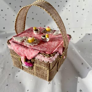 May include: A miniature picnic basket with a pink and white tablecloth, featuring miniature cupcakes and teacups. The basket is made of woven brown burlap and has a handle.