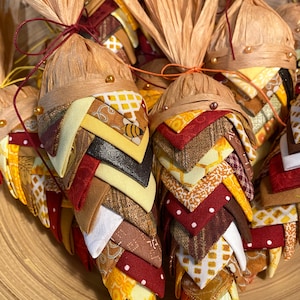 May include: A group of handmade fabric cornucopias with a fall theme. The cornucopias are made of various fabrics in shades of brown, red, yellow, and white. They are tied with twine and have a rustic look.