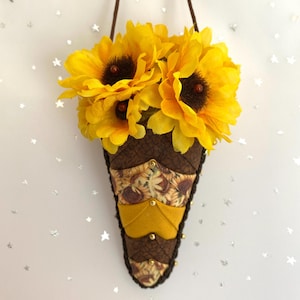 May include: A wall-hanging fabric cone filled with bright yellow artificial sunflowers. The cone features a layered, quilted design in shades of brown and mustard yellow, accented with small gold-toned studs and a dark brown braided trim edge.