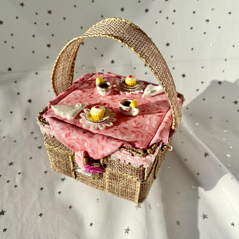 May include: A miniature picnic basket with a pink tablecloth and a handle made of burlap. The basket is filled with miniature teacups, saucers, and cakes.