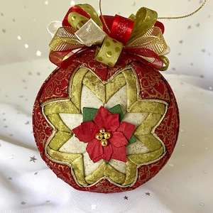 May include: A spherical fabric ornament featuring a quilted star pattern in red, gold, and cream fabric. The center is adorned with a red poinsettia flower with small gold beads at its core. A multi-layered bow in red and gold ribbon sits on top.