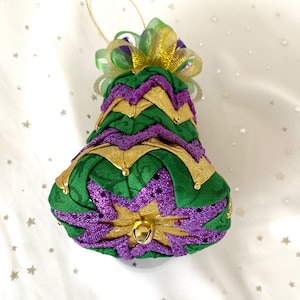 May include: A bell-shaped ornament in green, gold, and purple, with a gold bell at the bottom. The ornament has a gold string for hanging and is decorated with a green and gold bow.