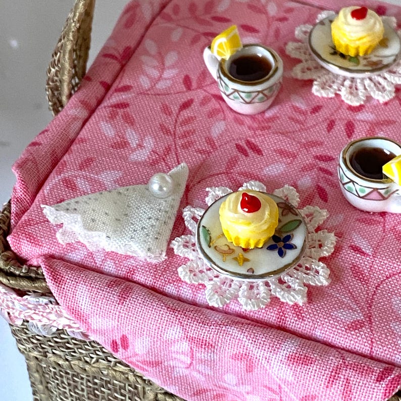 May include: A miniature wicker picnic basket with a pink floral tablecloth. The tablecloth has a white lace doily with a yellow cupcake with a red cherry on top. There are two teacups with lemon slices and tea, and a white doily with a pearl.