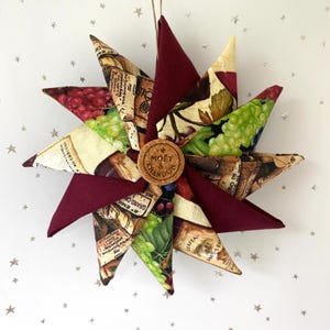 May include: A star-shaped ornament with alternating burgundy and patterned fabric panels. The patterned fabric features wine labels and grape illustrations. A cork with "Moët & Chandon" is in the center. A string is attached for hanging.