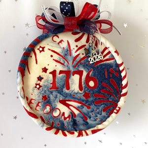 May include: A round patriotic ornament with a red, white, and blue design. The ornament features the number "1776" in red, along with the word "FREEDOM". It is decorated with a bow and a charm that says "2026".