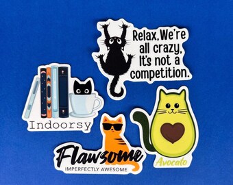 May include: A collection of cat-themed stickers on a blue background. The stickers feature various cat designs, including a black cat, an orange cat wearing sunglasses, and a cat inside a teacup. Text includes "Relax, We're all crazy, It's not a competition."