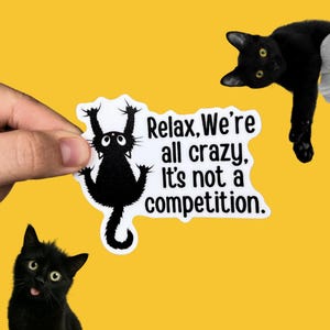 May include: Black and white sticker with a cartoon cat hanging upside down and the text "Relax, We're all crazy, It's not a competition."