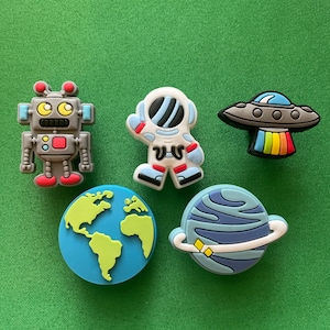 May include: Five colorful space-themed shoe charms: a robot, an astronaut, a flying saucer, a planet with rings, and a globe.