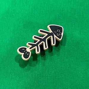 May include: A black and white fish bone charm with a cross on the tail fin. The charm is made of a beige rubber-like material.