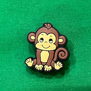 May include: A brown and yellow cartoon monkey charm with a curled tail. The monkey is sitting with its legs crossed and has a happy expression on its face.