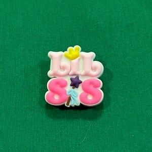 May include: A colorful shoe charm with the words "Lil Sis" in pink and white. The charm features a yellow hand, a purple star, and a small figure holding a blue staff. The charm is set against a green background.