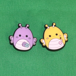 May include: Two cartoon-style character charms, one purple with flowers and one yellow with hearts. Both have smiling faces and are shaped like bugs.