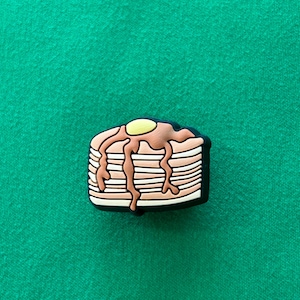 May include: A brown and white pancake charm with a yellow and brown syrup drizzle. The charm is made of rubber and has a black outline.