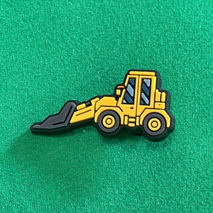 May include: A yellow and black bulldozer charm with a black scoop and wheels. The construction vehicle has a detailed cabin with blue-tinted windows. The charm is set against a green background.