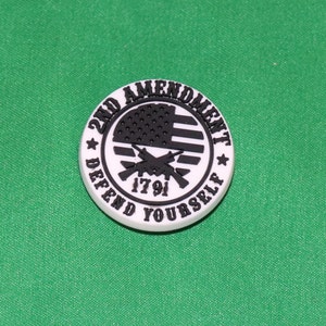 May include: A white round button with black text and a black and white American flag. The text reads "2nd Amendment Defend Yourself 1791".