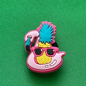 May include: A pink flamingo wearing sunglasses and a pineapple hat. The pineapple has yellow and green details. The flamingo is a charm or a decoration.
