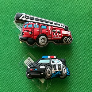 May include: Two toy vehicle charms, a red fire truck with a ladder and a black police car with a blue light. Both charms have a cartoon style and are made of rubber.