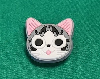May include: A gray and white cartoon cat charm with pink ears. The cat has black eyes and a pink tongue sticking out.