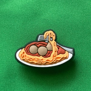 May include: A colorful shoe charm featuring a plate of spaghetti and meatballs. The charm has a black border and includes a fork with spaghetti. The meatballs are brown, and the sauce is red.
