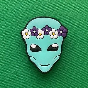 May include: A light blue alien face with a white and purple flower crown. The alien has black eyes and a small smile.