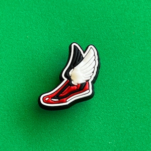 May include: A red, white, and black shoe charm with a wing on the back. The shoe is red with a white sole and a black wing. The wing is white with a black outline.
