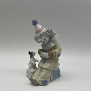 May include: A porcelain figurine of a clown playing an accordion with a small dog sitting on a dice block. The clown is wearing a colourful costume with a blue and white hat.
