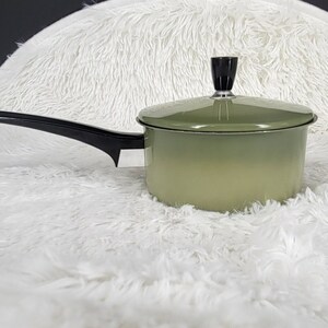 May include: A vintage green saucepan with a black handle and lid. The saucepan has a rounded body and a long, angled handle. The lid has a black knob. The pan is set against a white, fluffy background.