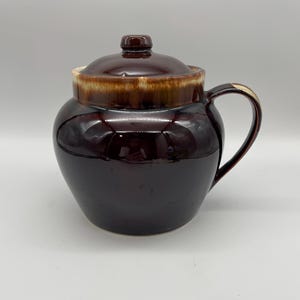 May include: A dark brown ceramic crock with a lid and handle. The crock has a glossy finish and a lighter brown band around the rim. The lid has a small knob on top.