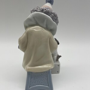 May include: A porcelain figurine of a clown with a blue and white hat, wearing a beige jacket and sitting on a blue stool. The clown is holding a small white dog.