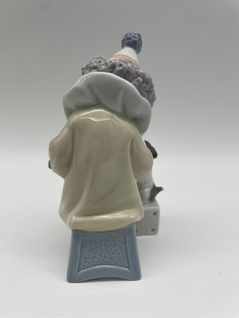 May include: A porcelain figurine of a clown with a blue and white hat, wearing a beige jacket and sitting on a blue stool. The clown is holding a small white dog.