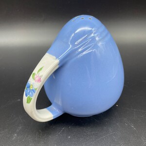 May include: A blue ceramic salt shaker with a white handle and floral design. The shaker has a round shape and a small opening on the top for pouring.