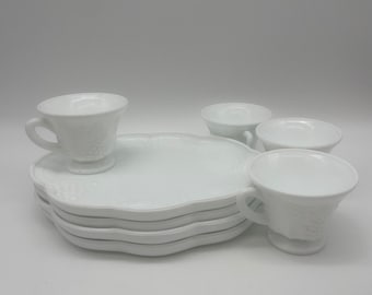May include: A set of white milk glass dinnerware featuring a stack of four scalloped plates and three matching footed teacups. Each piece is decorated with a subtle embossed grape and vine pattern along the edges, creating a uniform look.