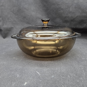 May include: A vintage, amber-colored glass casserole dish with a matching lid. The dish has a rounded shape with two small handles and a knob on the lid. The glass has a reflective surface.
