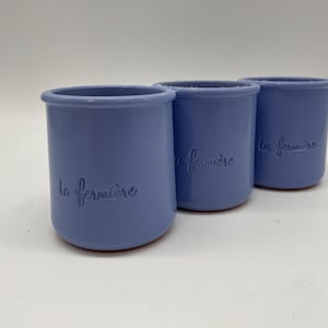 May include: Three light blue ceramic jars with the text "la fermière" embossed on the side. The jars have a cylindrical shape with a slightly flared rim and a reddish-brown base. The jars are arranged in a row against a white background.