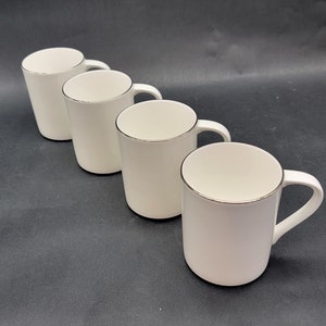 May include: Four white ceramic mugs with silver rims and white handles are arranged diagonally on a dark surface. The mugs have a simple, elegant design, suitable for coffee or tea. The mugs are evenly spaced and appear to be new.