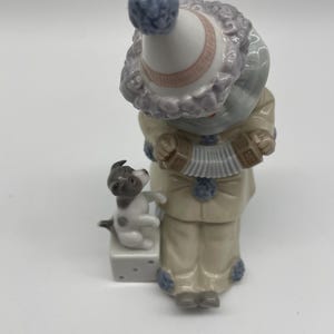 May include: A porcelain figurine of a clown playing an accordion with a small dog sitting on a dice block. The clown is wearing a blue and white hat with a pink band, a blue and white striped shirt, and beige trousers. The dog is white with black spots.