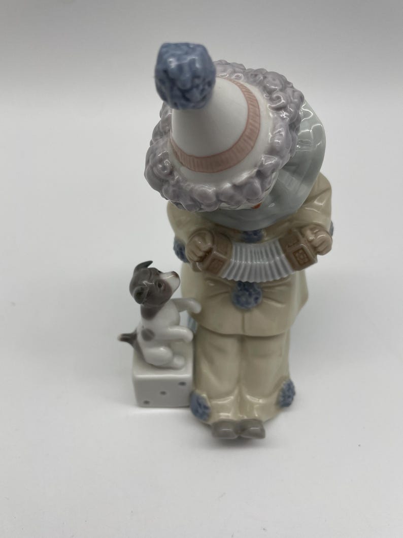 May include: A porcelain figurine of a clown playing an accordion with a small dog sitting on a dice block. The clown is wearing a blue and white hat with a pink band, a blue and white striped shirt, and beige trousers. The dog is white with black spots.