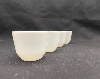 May include: Four white ceramic cups are arranged in a row, each with a rounded base and a slightly flared rim. The cups have a smooth, matte finish and are set against a dark background.